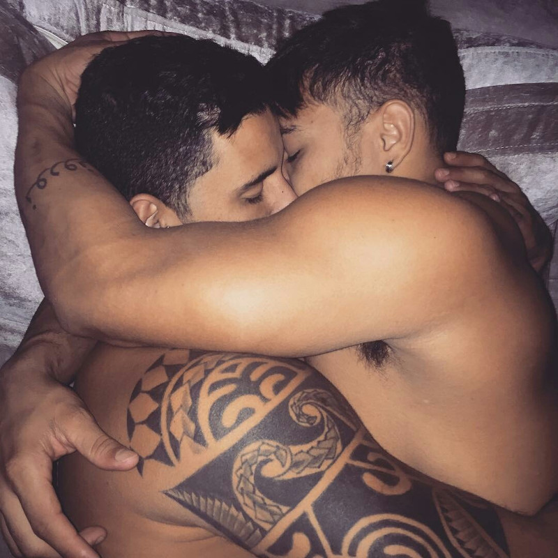 Two shirtless, tattooed men lying in bed, hugging each other tenderly.