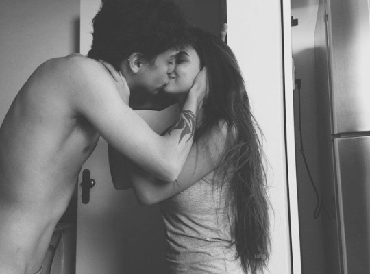 A black and white photo of a guy and a girl kissing in the kitchen doorway.