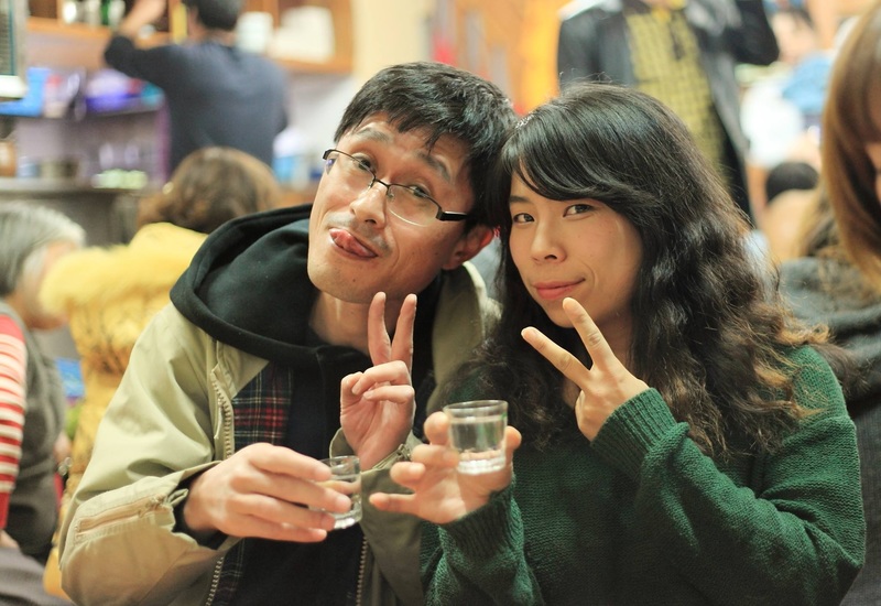 A man and a woman in a bar, each holding a shot glass with liquor. They are posing for the camera, making peace signs. The guy is sticking his tongue out.