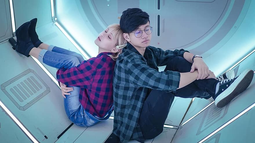 A young woman and a young man are sitting back to back on the floor of a room that looks like an airlock. They're both wearing plaid shirts and jeans, and have their eyes closed. Are they dating?