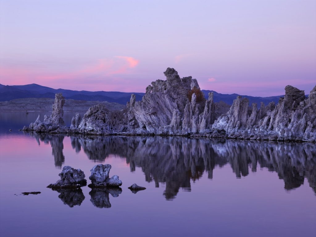 An outcropping of eroded rock formations advancing into the calm waters of a lake. It's sunset and the sky has hues of the bisexual pride colours, pink, purple and blue. The water reflects the sky's pretty colours. The image is about the calm that radical acceptance can bring, which helps dealing with internalized biphobia and homophobia.