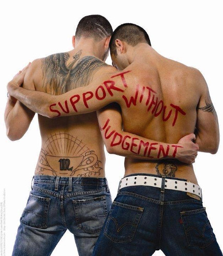 A view from behind of two shirtless men side by side, embracing one another. The words "support with judgement" are painted in red on their exposed skin. Both men have multiple tattoos, buzzed hair, and are wearing jeans. This kind of support is essential to heal from internalized biphobia and homophobia