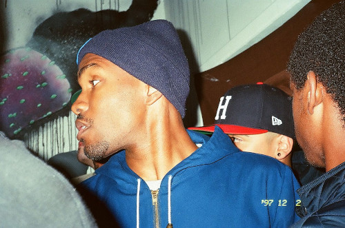 Frank Ocean, a Black singer and rapper who is openly bisexual. He is standing among a crowd indoors. He's wearing a hoodie and a beanie hat.