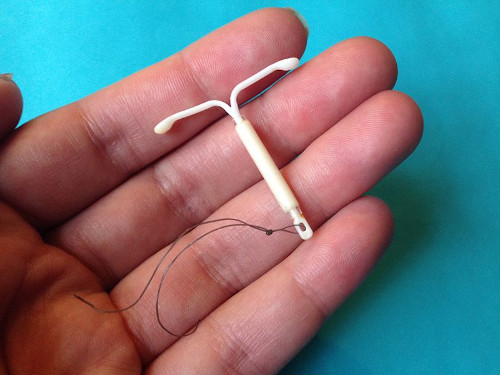 A plastic intra-uterine device resting in someone's fingers. It is T-shaped, and it has a wire tied to the bottom end of it. It is barely longer than the width of two fingers.